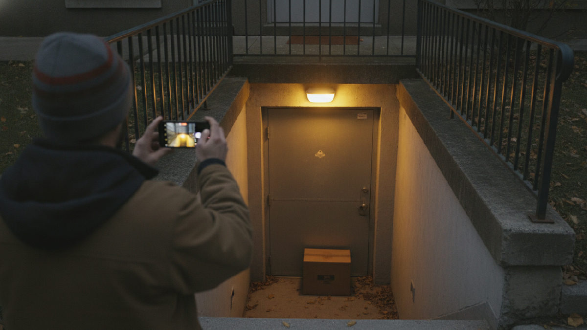 Employé vu de dos, tuque et manteau de travail, qui photographie avec son cellulaire une boîte déposée devant une porte de service au bas d'un escalier de sous-sol couvert de feuilles mortes, un soir d'automne